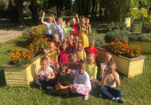 w ogrodzie dzieci w kolorowych ubraniach pozują zajmując miejsca przy żółtych skrzynkach z pomarańczowymi, jesiennymi kwiatami